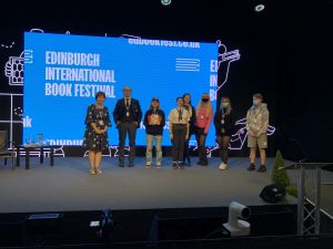 Image shows Prof Stella Chan and the young citizen scientists on stage at the book festival in front of a blue screen that says 'Edinburgh International Book Festival'